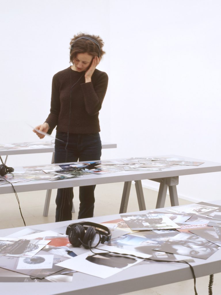 Photograph of the PLOTS installation showing one person standing at a table using the headphones to listen to the installation audio.