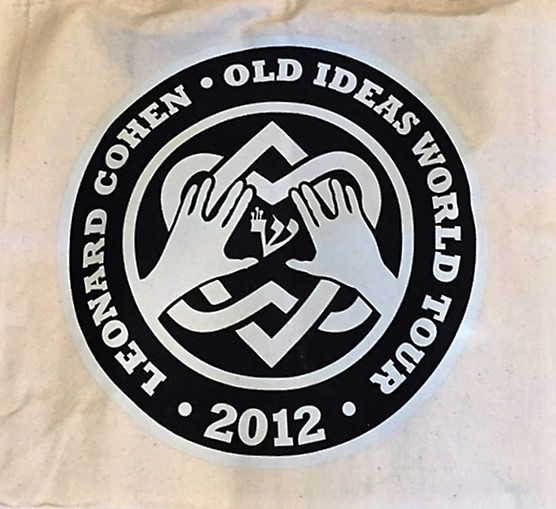A circular logo on a white background. The circle is black with white text around the rim reading "Leonard Cohen Old Ideas world Tour 2012". The interior of the cirlce contains a white design of two interlocking hearts and two hands.